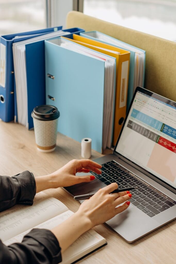 a-person-using-a-laptop-8296985 Hands typing on a laptop at an office desk with files and coffee.
