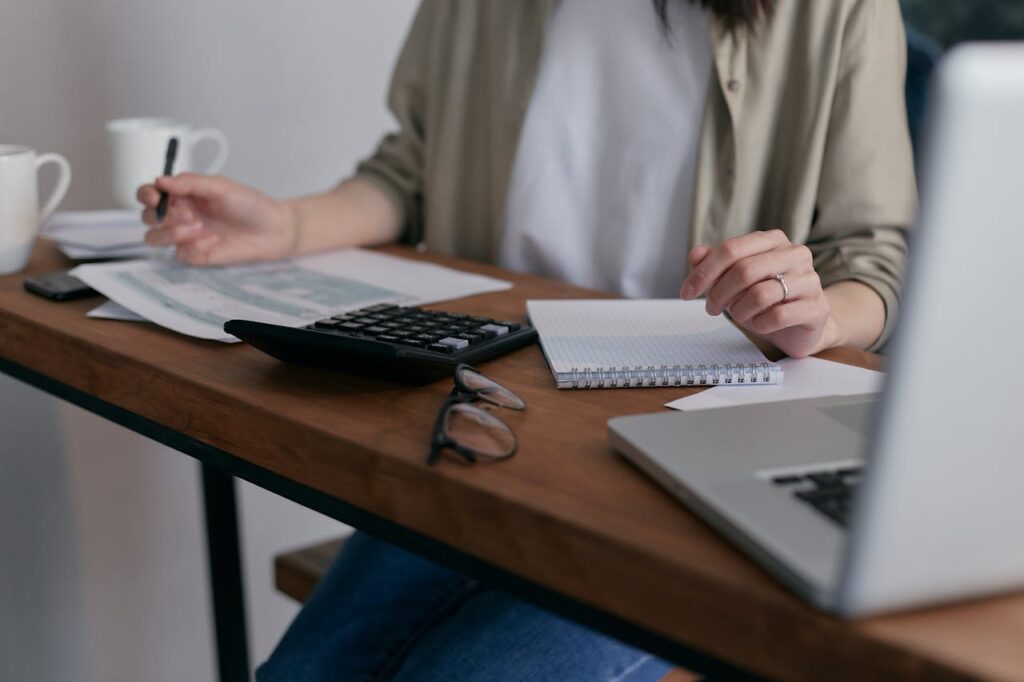 couple-people-woman-desk-6963857 A woman manages finances at home, using a laptop and calculator on a wooden desk.