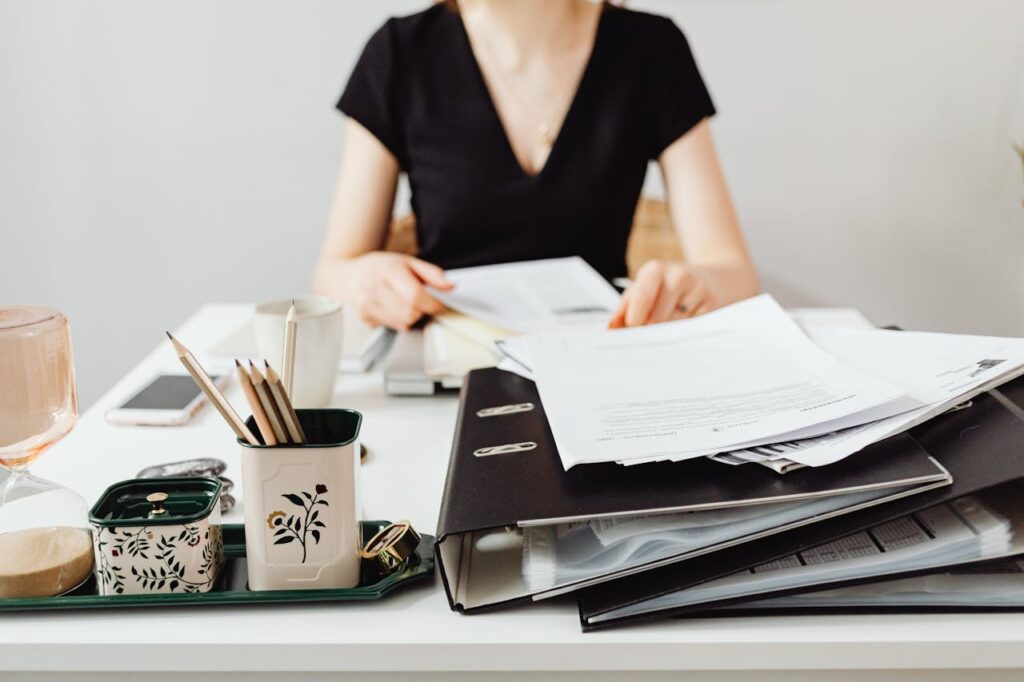 documents-on-top-of-a-desk-7681332 A woman at a desk working with binders and documents, featuring essential office supplies.