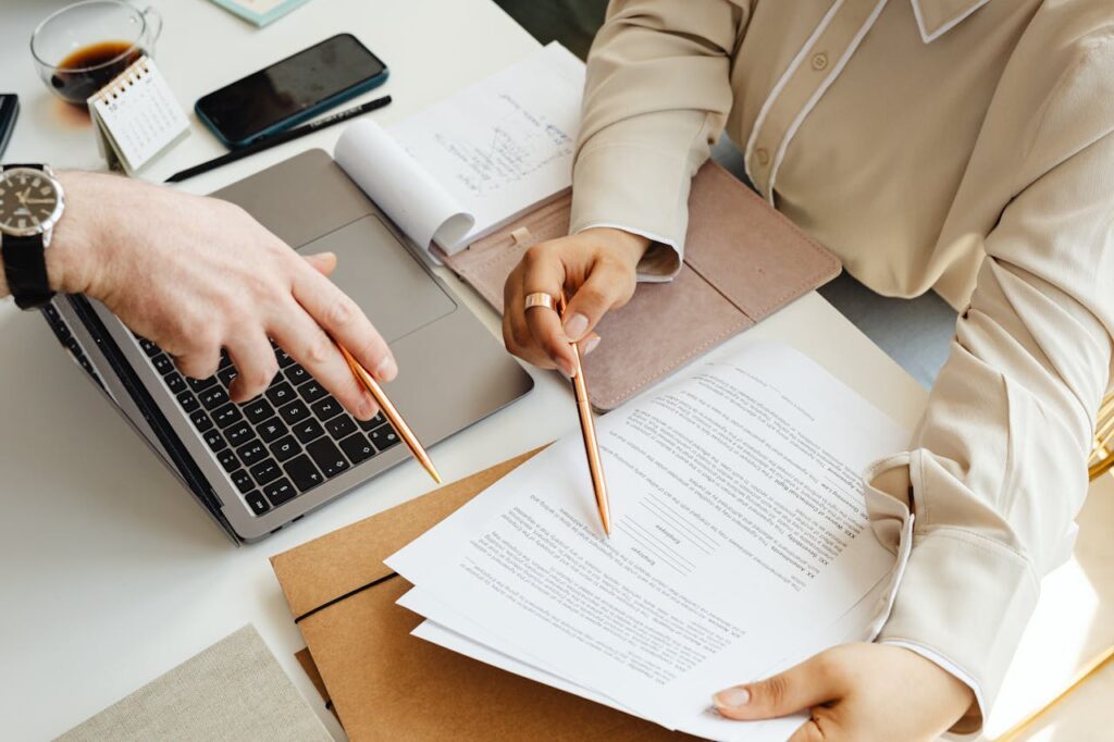 people-pointing-their-pens-on-a-printed-paper-7876785 Office teamwork with two people reviewing documents and using a laptop