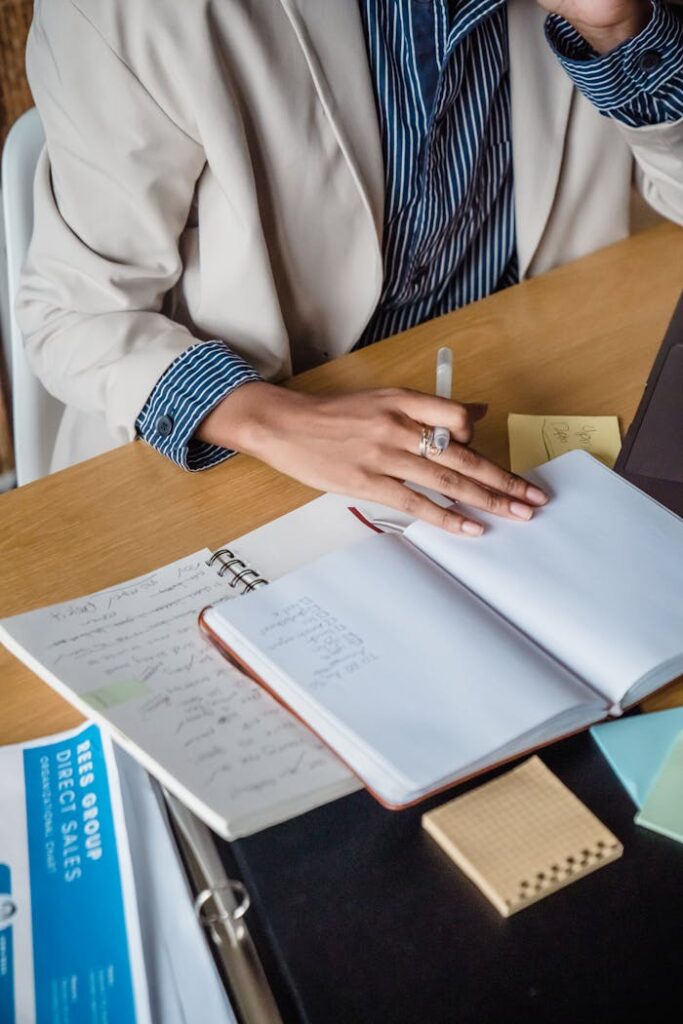 woman-sitting-behind-a-desk-and-writing-in-a-notebook-12912110 Woman writing in a notebook at a desk with papers and sticky notes, highlighting office productivity.